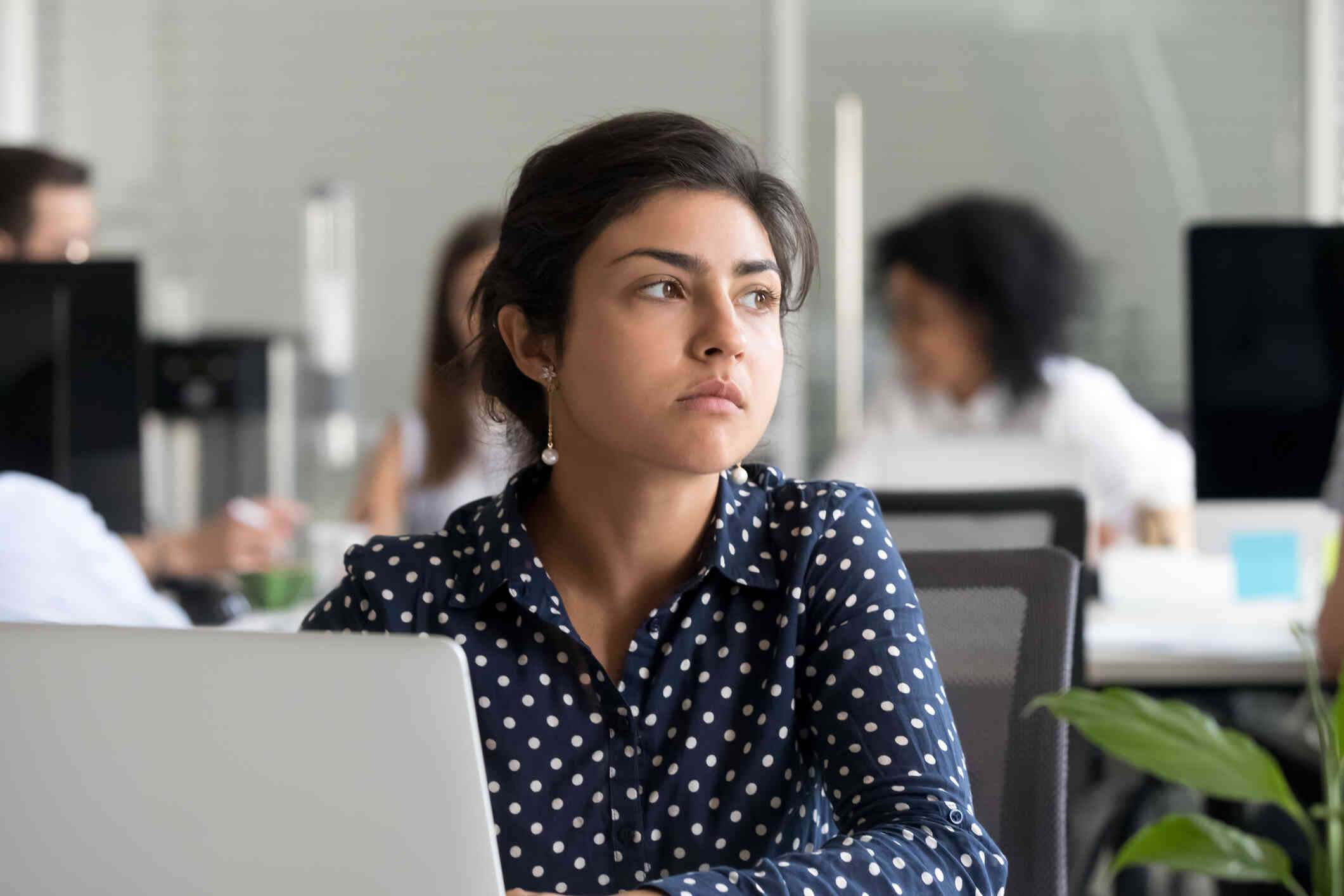 A woman sits at her desk at work behind her computer and gazes off with a sad expression.